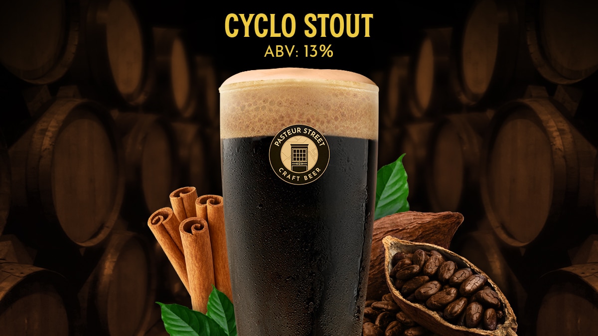 A glass of Pasteur Street Cyclo Stout, a dark stout with its key ingredients, cinnamon sticks and a split cacao pod, in front of aged wooden barrels.
