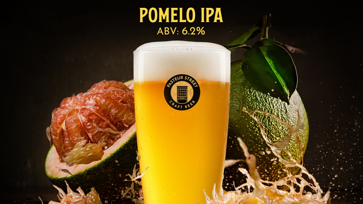 A glass of Pasteur Street Pomelo IPA with a peeled pomelo showing its pink flesh and a whole green pomelo with a liquid splash.